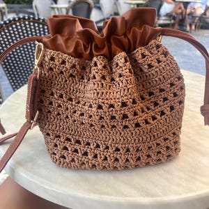 K&ouml;nnte beinhalten: Eine braune Beuteltasche mit einem geflochtenen, stroh&auml;hnlichen K&ouml;rper in einem offenen Strickmuster und einem glatten, braunen Kordelzugverschluss aus Kunstleder. Die Tasche verf&uuml;gt &uuml;ber einen passenden Schulterriemen aus Kunstleder.