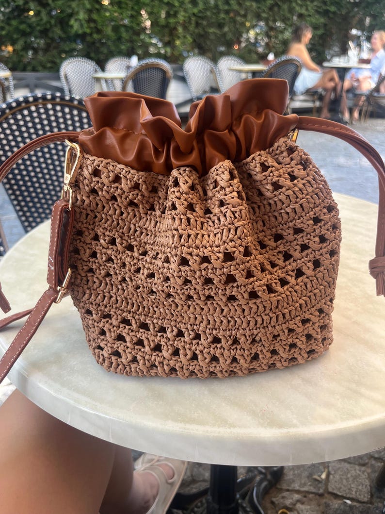 K&ouml;nnte beinhalten: Eine braune Beuteltasche mit einem geflochtenen, stroh&auml;hnlichen K&ouml;rper in einem offenen Strickmuster und einem glatten, braunen Kordelzugverschluss aus Kunstleder. Die Tasche verf&uuml;gt &uuml;ber einen passenden Schulterriemen aus Kunstleder.