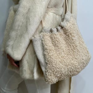 May include: A cream-colored, textured shoulder bag with a drawstring closure. The bag has a fluffy, shaggy exterior and a neutral-toned, woven fabric at the top. The bag is carried by a cream-colored cord.