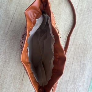 K&ouml;nnte beinhalten: Eine Beuteltasche mit geflochtenem Stroh-Au&szlig;enmaterial und glattem, braunem Kunstlederbesatz. Die Tasche verf&uuml;gt &uuml;ber einen Kordelzug, einen langen, d&uuml;nnen Schulterriemen und ein wei&szlig;es Stofffutter im offenen Hauptfach.