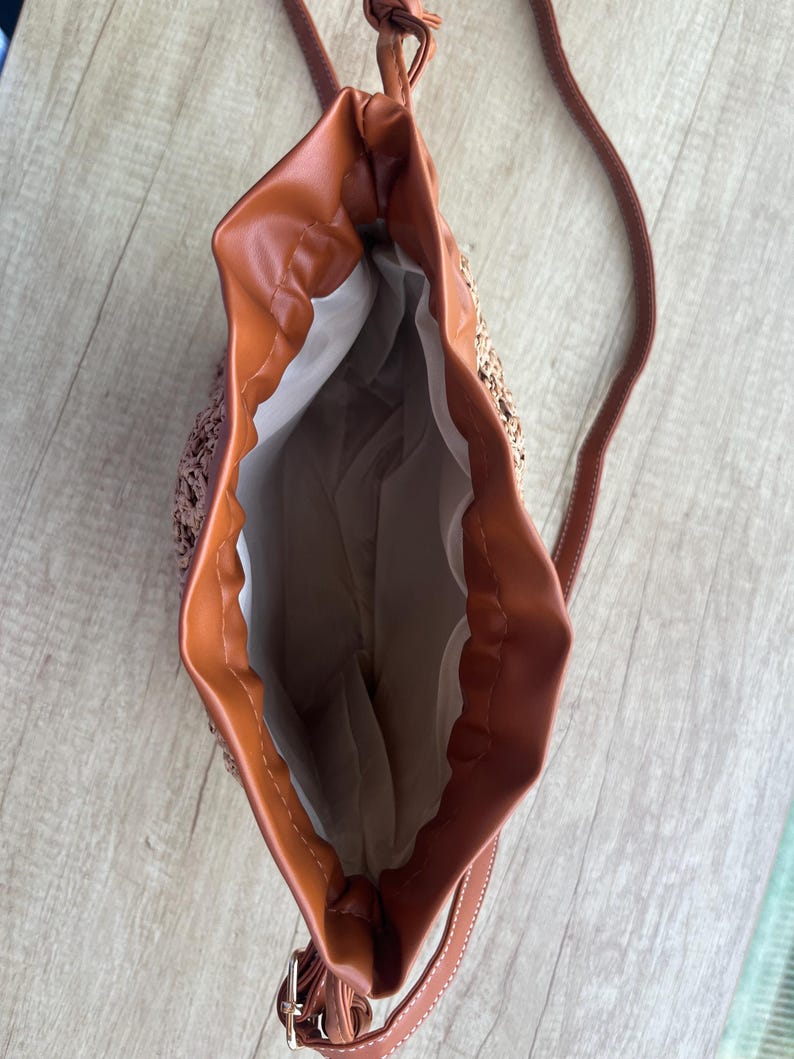 K&ouml;nnte beinhalten: Eine Beuteltasche mit geflochtenem Stroh-Au&szlig;enmaterial und glattem, braunem Kunstlederbesatz. Die Tasche verf&uuml;gt &uuml;ber einen Kordelzug, einen langen, d&uuml;nnen Schulterriemen und ein wei&szlig;es Stofffutter im offenen Hauptfach.