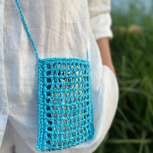 May include: A turquoise crochet phone bag with a long strap. The open weave design allows a glimpse of the phone inside. The bag is worn over a white shirt, suggesting a casual, summery style. The bag is a vibrant accessory.