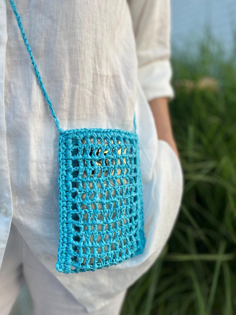 May include: A turquoise crochet phone bag with a long strap. The open weave design allows a glimpse of the phone inside. The bag is worn over a white shirt, suggesting a casual, summery style. The bag is a vibrant accessory.