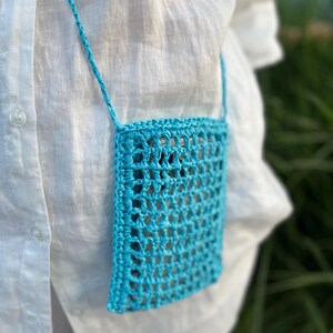 May include: A turquoise crochet crossbody bag with a woven design. The bag is rectangular and has a matching strap. The background is a white shirt and green foliage.