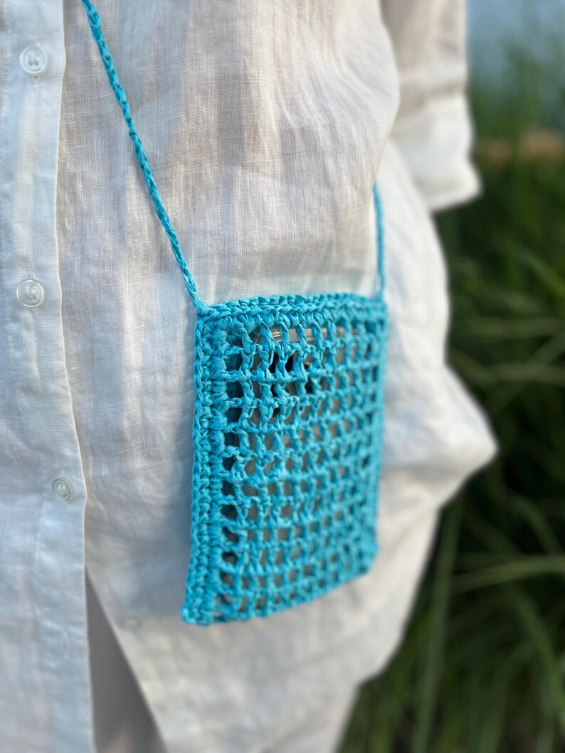 May include: A turquoise crochet crossbody bag with a woven design. The bag is rectangular and has a matching strap. The background is a white shirt and green foliage.
