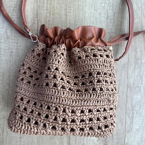 K&ouml;nnte beinhalten: Eine beigefarbene H&auml;kel-Beuteltasche mit Kordelzug und strukturiertem, offenem Webmuster. Die Tasche verf&uuml;gt &uuml;ber einen glatten, braunen Kunstleder-Oberteil mit geraubtem Kordelzugverschluss und einen passenden d&uuml;nnen braunen Schulterriemen mit wei&szlig;en N&auml;hten.
