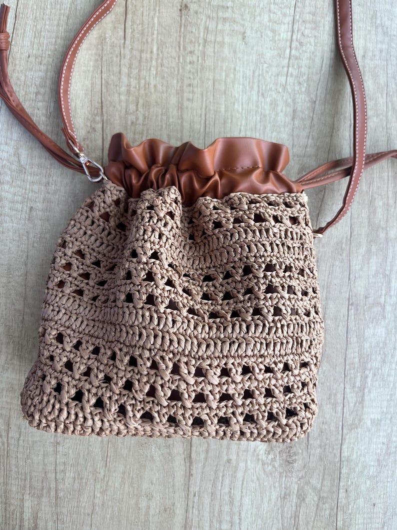K&ouml;nnte beinhalten: Eine beigefarbene H&auml;kel-Beuteltasche mit Kordelzug und strukturiertem, offenem Webmuster. Die Tasche verf&uuml;gt &uuml;ber einen glatten, braunen Kunstleder-Oberteil mit geraubtem Kordelzugverschluss und einen passenden d&uuml;nnen braunen Schulterriemen mit wei&szlig;en N&auml;hten.