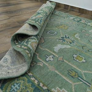 Op de afbeelding: Een gedeeltelijk opgerold, rechthoekig tapijt met een lichtgroene achtergrond en ingewikkelde bloempatronen in tinten groen, blauw en beige. De rand van het tapijt heeft een gedetailleerd geometrisch ontwerp. Het tapijt wordt op een houten vloer getoond.