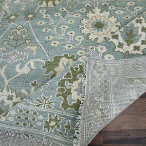 Op de afbeelding: Een close-up van een blauw en groen patroonkleed met een bloemenontwerp. Het kleed heeft een versleten uitstraling, met licht gerafelde randen. Het kleed is opgevouwen, waardoor de onderkant zichtbaar is, en ligt op een houten oppervlak.