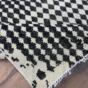May include: A black and cream-colored rug with a repeating checkered pattern. The rug is partially rolled up, revealing the fringed edge. The design is a classic, geometric pattern, suitable for various interior styles. The rug appears to be made of a woven material.