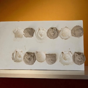 以下が含まれることがあります： 白の長方形の型に、グレーと白の様々な色合いの猫の顔の印象が12個あります。猫の顔は様々なスタイルで、詳細な特徴を持つものもあれば、より抽象的なものもあります。