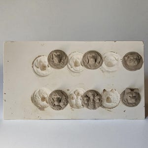 以下が含まれることがあります： 様々な犬種が円形に型取られた、オフホワイトの長方形の型。犬の顔は白と茶色の色合いで、詳細な顔の特徴が示されています。型は石膏または同様の素材で作られているようです。