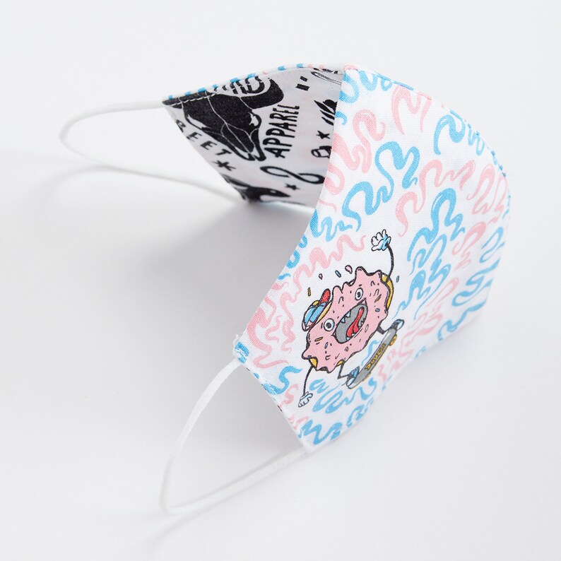May include: White face mask with a pink and blue donut character skateboarding on a white background with blue and pink swirls. The text "Donut Apparel" is visible on the other side of the mask.
