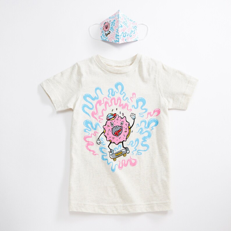 May include: A light beige t-shirt with a pink and blue donut character skateboarding. The donut character is wearing a blue hat and has a pink and white frosting design. The donut character is surrounded by pink and blue swirls. The t-shirt is paired with a face mask with a pink and blue donut pattern.