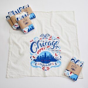 May include: Set of four white tea towels with a blue and red design featuring the text "Chicago" and the skyline of the city. The text "City by the Lake" and "EST. 1833" are also included in the design.