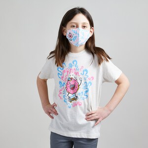 May include: A young person wearing a white t-shirt with a pink and blue donut character graphic. The donut character is skateboarding and surrounded by blue and pink swirls. The person is also wearing a pink and blue patterned face mask.
