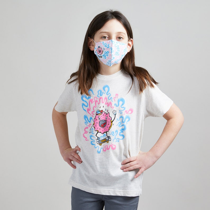 May include: A young person wearing a white t-shirt with a pink and blue donut character graphic. The donut character is skateboarding and surrounded by blue and pink swirls. The person is also wearing a pink and blue patterned face mask.