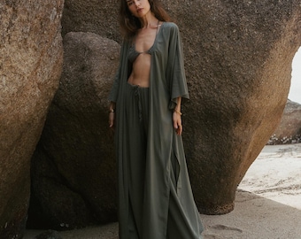 Może przedstawiać: Oliwkowy zestaw składający się z topu od bikini, spodni z wysokim stanem i długiej kurtki w stylu kimono. Strój wykonany jest z lekkiego materiału. Modelka stoi na piaszczystej plaży z dużym formacją skalną w tle.