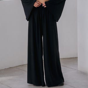 May include: A black two-piece outfit featuring a long-sleeved crop top with a plunging neckline and a large gold ring detail. The outfit includes wide-leg pants. The model is wearing a necklace and hoop earrings.