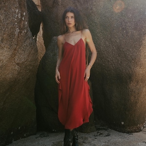 May include: A woman in a bright red slip dress with thin straps and black boots. The dress has a flowing design. The background shows large, textured rock formations, with sunlight creating a warm effect. The dress is approximately 150 cm in length.