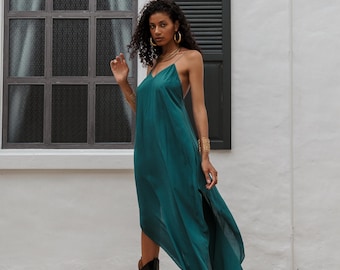 May include: A teal green, flowing maxi dress with spaghetti straps and a high side slit. The dress is paired with black cowboy boots and gold jewelry. The model is standing in front of a white wall with a window and black shutters.