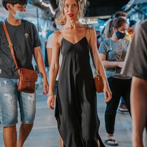 Pode incluir: Uma mulher em um vestido preto com alças finas caminha por um mercado ao ar livre lotado. Ela está com o cabelo preso e usando batom vermelho. Outras pessoas estão ao fundo, algumas usando máscaras.
