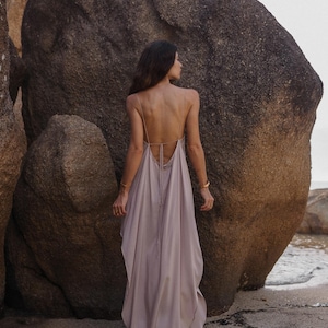 May include: A long, flowing dress in a pale mauve colour, featuring delicate straps and a low back. The dress falls to the ground, creating a soft, elegant shape. The model is on a sandy beach with large rocks in the background.