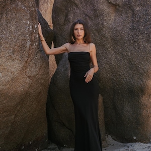 May include: A long, black dress with thin straps. The dress has a fitted top and a flowing skirt. The model is wearing gold hoop earrings. The background features large rock formations and sand.