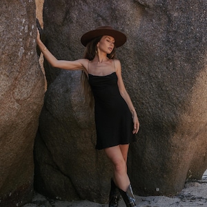 May include: A woman in a black slip dress, brown hat, and black cowboy boots stands by large rocks. The dress has thin straps and a draped neckline. The boots are knee-high.
