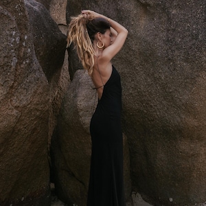 May include: A woman in a black backless maxi dress stands near large rocks. She has blonde hair pulled back and is wearing gold hoop earrings. The dress has thin straps and a low back, and the skirt flows to the ground.