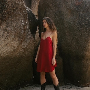 May include: A person wearing a red slip dress and black cowboy boots stands between large rock formations. The dress has thin straps and a V-neck. The boots are black with decorative stitching.