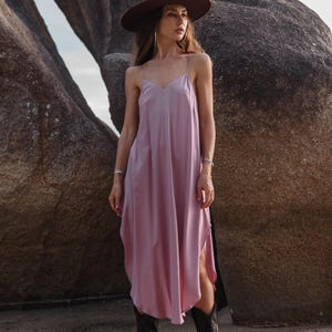May include: A mauve slip dress with spaghetti straps, a brown wide-brimmed hat, and black cowboy boots. The dress has a V-neck and a side slit. The boots have a pointed toe and decorative stitching. The outfit is worn outdoors.