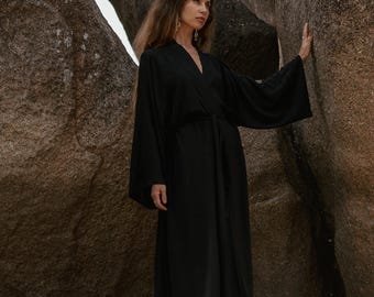 May include: A long, black kimono-style robe with wide sleeves and a matching belt. The robe is worn over white pants. The model is standing in front of a large rock formation.