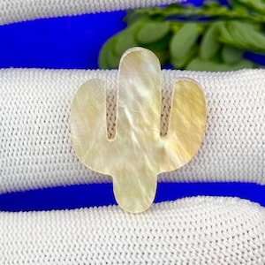 May include: A pearlescent, cactus-shaped pendant with a natural, iridescent finish. The pendant is set against a blue background and rests on a white, textured surface.