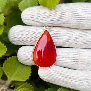 May include: A teardrop-shaped pendant with a vibrant red gemstone set in a silver-coloured bezel. The polished stone reflects light, creating a glossy appearance. The pendant is displayed against a white background.