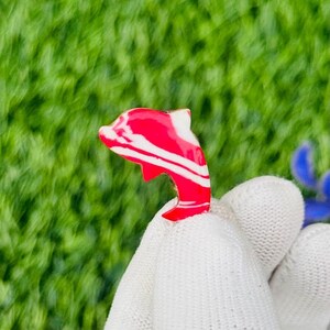 Op de afbeelding: Een rood en wit dolfijnvormig bedeltje. De bedel heeft een wervelpatroon van rood en wit. De dolfijn wordt vastgehouden door een witte handschoen tegen een groene achtergrond.