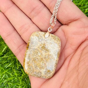 Könnte beinhalten: Eine rechteckige Anhängerhalskette mit einer silbernen Kette. Der Anhänger ist ein hellbrauner Stein mit einem einzigartigen, strukturierten Muster. Der Stein hat eine polierte, glänzende Oberfläche und wird von einer silbernen Öse gehalten.