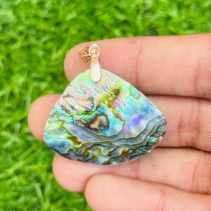 May include: Abalone shell pendant with iridescent blues, greens, and purples. The pendant is triangular with a gold-colored bail. The natural shell has a textured, layered appearance, showcasing the unique patterns of the abalone.