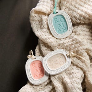 Puede incluir: Tres etiquetas ovaladas blancas para el hogar con la palabra "ROMAN" en el centro. Las etiquetas tienen centros de diferentes colores: azul claro, melocotón y crema. Cada etiqueta está unida a una cuerda con una pequeña perla.