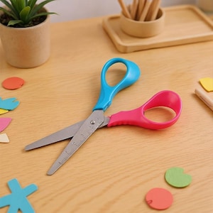 May include: A pair of scissors with a blue handle and a pink handle. The blades are silver and have a ruler printed on them. The scissors are open and resting on a light brown wooden surface. Colorful craft foam shapes are scattered around the scissors.