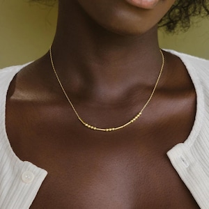 Puede incluir: Un delicado collar dorado con una serie de pequeñas cuentas y barras cilíndricas. El collar se lleva alrededor del cuello. Es una joya sencilla y elegante. El collar es de oro.
