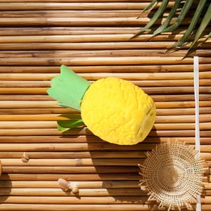 May include: A yellow pineapple with green leaves rests on a bamboo mat. A white straw and a small woven coaster are also visible. The tropical fruit is a bright, sunny yellow, and the leaves are a vibrant green.
