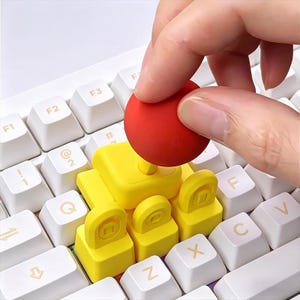 May include: A close-up of a white keyboard with yellow and red keycaps. The yellow keycaps are square with a rounded top and a small square in the center. A red ball-shaped keycap is being placed on the keyboard.