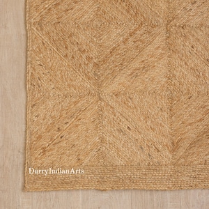 May include: Close-up of a natural jute rug with a geometric diamond pattern. The rug is a warm tan color and has a textured surface. The edge of the rug has a braided border. The text "DurryIndianArts" is visible.