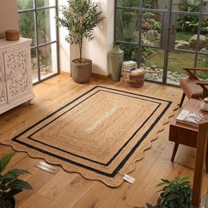 May include: A rectangular jute rug with a scalloped edge and a black border. The rug is light brown with a black double border and the text "DhurriesIndiaArt" in white. The rug is placed on a wooden floor.