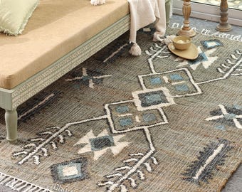 May include: A rectangular, gray rug with a geometric pattern in white, blue, and black. The rug has a fringe on the edge and is placed near a beige bench. The rug's design includes diamond and star shapes.