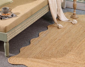 May include: A natural jute rug with a scalloped edge is shown on a grey surface. The rug is light tan, with a woven texture. The rug is near a beige daybed with a decorative border and a white throw blanket.