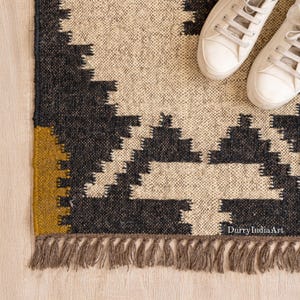May include: A woven rug with a geometric pattern in black, beige, and mustard yellow. The rug has a fringed edge and is paired with white canvas trainers. The rug's design features repeating triangular shapes.