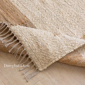 May include: A natural fibre rug with a textured, woven design. The rug has a cream-coloured central section and a brown border. The edge of the rug is fringed. The text "DurryIndiaArts" is visible in the lower left corner.