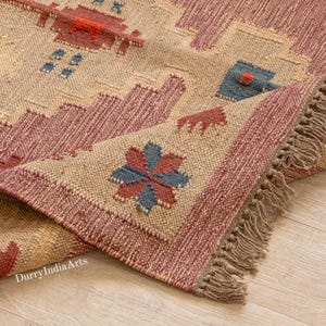 May include: Close-up of a beige and burgundy woven rug with a geometric pattern. The rug features a repeating design with burgundy and blue accents, and a fringed edge. The text "DurryIndiaArts" is visible.
